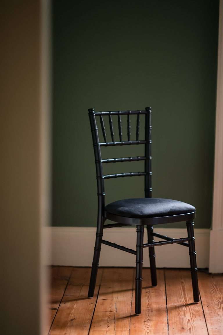 A black chiavari chair with a black velvet seat pad taken against a dark green background